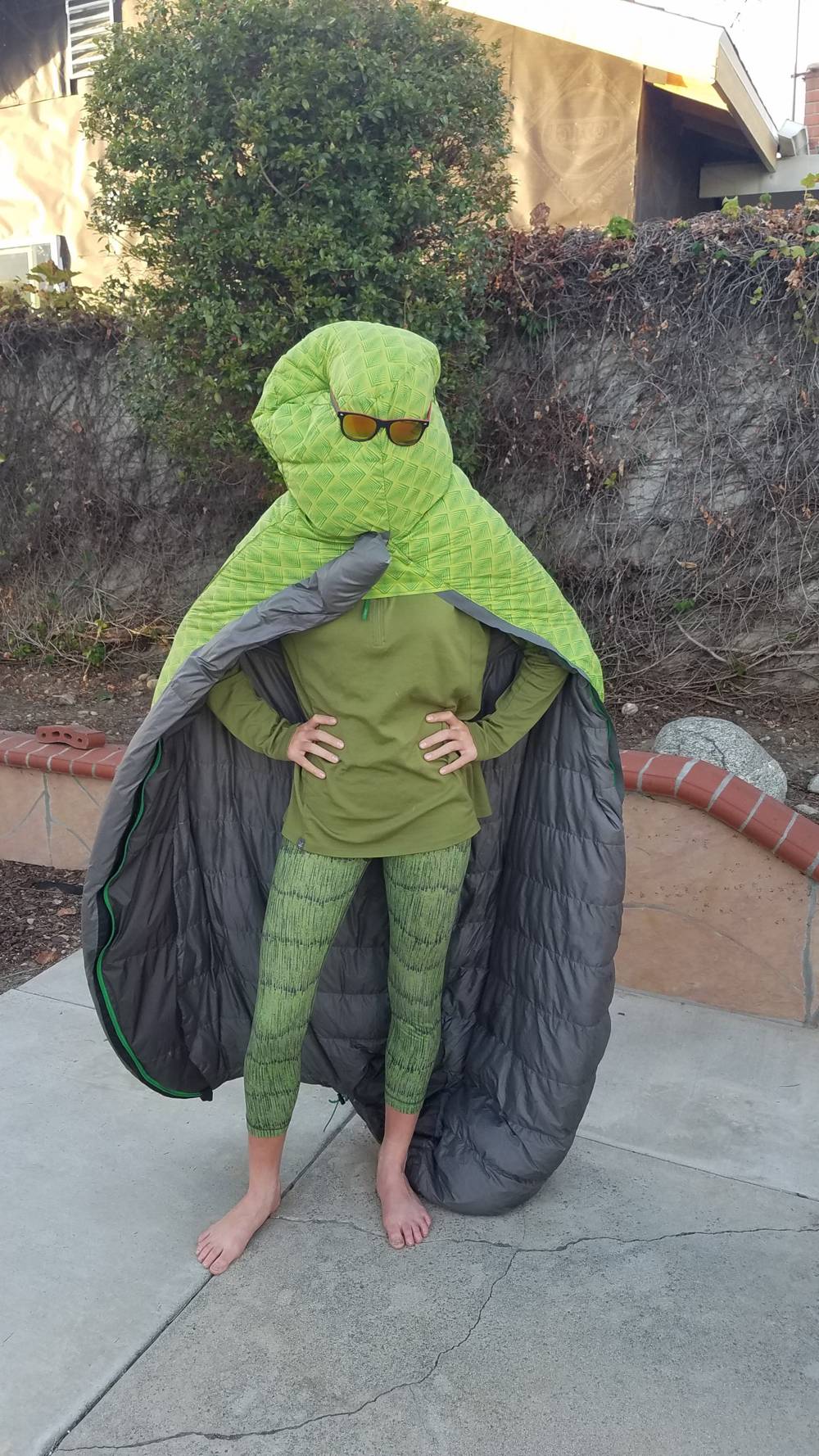 A barefoot child stands with hands on hips in green clothes, wearing a green patterned sleeping bag as a hood and cape. The bag’s gray lining is exposed. The hood covers the child’s face, with orange-lens sunglasses sitting on it.