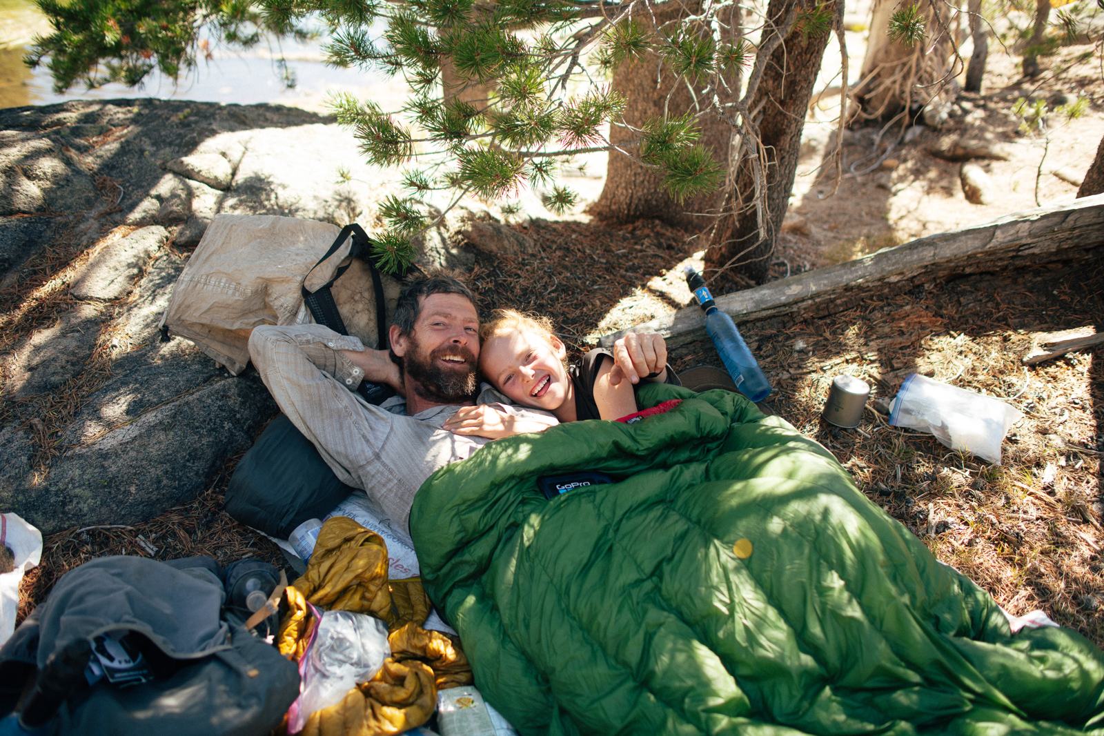 Father and child lie cheek to cheek beneath pine branches, both smiling; a blue bottle and metal cup sit beside them.