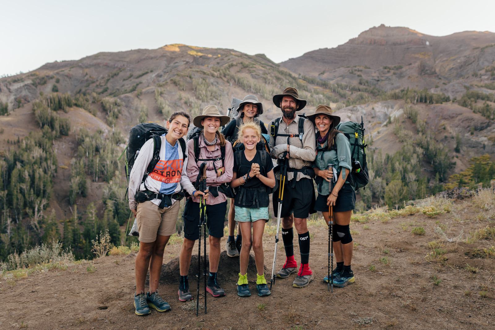 Six family members stand close together on a dirt ridge, smiling toward the camera, wearing backpacks and holding trekking poles.