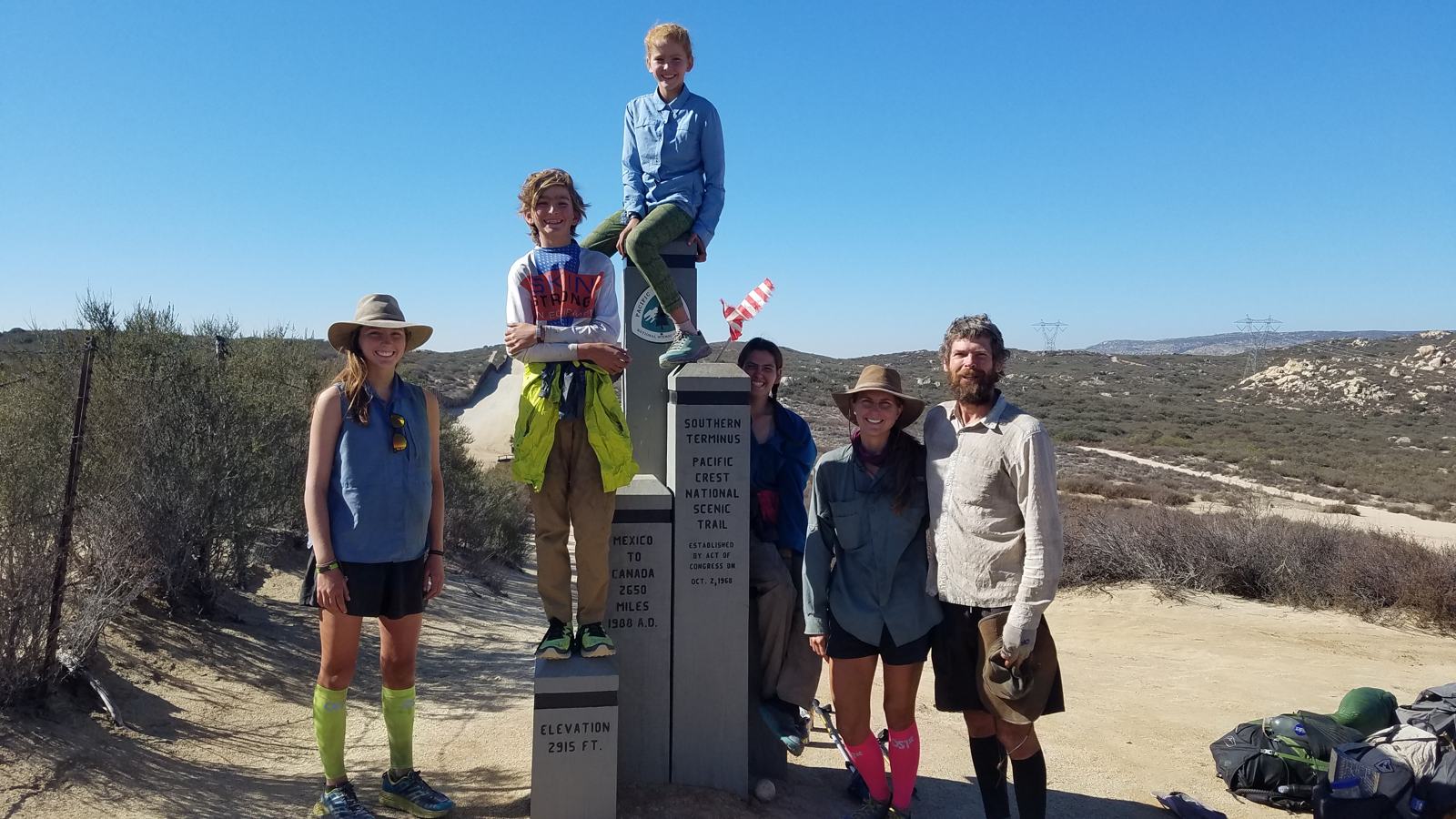 Six people pose around the monument, one child seated on its tallest post; a small striped flag and piled backpacks are visible.